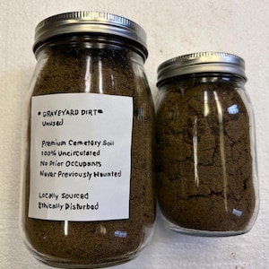 May include: Two glass jars filled with dark brown soil. The larger jar has a label that reads "GRAVEYARD DIRT Unused Premium Cemetery Soil 100% Uncirculated No Prior Occupants Never Previously Haunted Locally Sourced Ethically Disturbed."