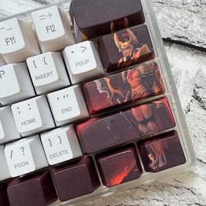 Pode incluir: Uma aproximação de um teclado mecânico com teclas personalizadas. As teclas são uma mistura de branco e vermelho escuro. Algumas teclas apresentam ilustrações de um guerreiro com armadura dourada e vermelha. O teclado está sobre uma superfície cinza claro.