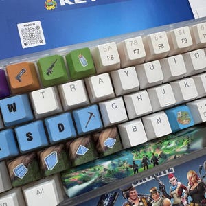 May include: A keyboard with custom keycaps featuring game-themed designs. The keycaps have various colors and include images of weapons, landscapes, and other game-related icons. The word "KEYCAPS" is visible in the background.