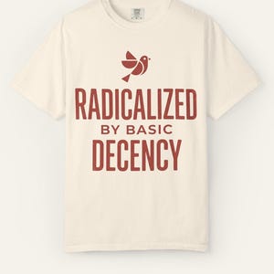 Puede incluir: Camiseta color crema con un gráfico de pájaro rojo y la frase "RADICALIZED BY BASIC DECENCY". El texto está en negrita. La camiseta tiene cuello redondo y parece estar hecha de un tejido suave y cómodo.