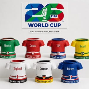 May include: A selection of ceramic shirt-shaped containers, each representing a country in the 2026 World Cup. The containers are in various colours, including green, red, blue, and yellow, with country names and logos. The FIFA World Cup logo is visible.