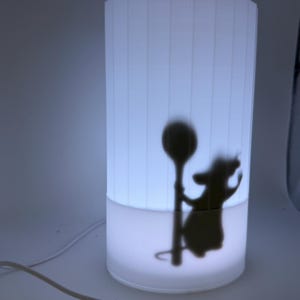 May include: A cylindrical white lamp featuring a silhouette of a rat holding a spoon. The lamp has a vertical striped pattern and emits a soft, diffused light. A white power cord is visible.