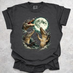 May include: Dark grey t-shirt with a graphic of three alligators and a full moon. The alligators have open mouths and raised arms, set against a starry night sky. The design is detailed and artistic.