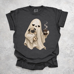 Pode incluir: Uma t-shirt cinzento-carvão de manga curta com um gráfico de um fantasma bege com uma expressão aborrecida, segurando um pug fulvo num braço e uma caneca de café castanho-escura com a palavra MEH e vapor na outra mão.