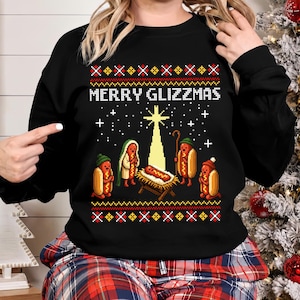 May include: A black long-sleeve sweatshirt featuring a pixel art graphic of a nativity scene with hot dogs. The text MERRY GLIZZMAS is printed above the scene, which includes a hot dog in a manger, a star, and hot dog figures in winter hats.