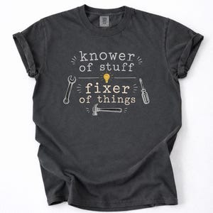 May include: A charcoal gray t-shirt featuring the white text "knower of stuff" and "fixer of things" with a yellow lightbulb icon. The design includes illustrations of a wrench, a screwdriver, and a hammer arranged around the text.
