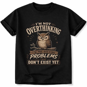 May include: A black short-sleeved t-shirt featuring a graphic of a brown owl perched on a branch. The text reads: I'M NOT OVERTHINKING I'M PRE-SOLVING PROBLEMS THAT DON'T EXIST YET. The design uses tan and brown tones against the black fabric.