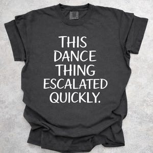 May include: Dark gray t-shirt with the text "THIS DANCE THING ESCALATED QUICKLY" in white. The shirt is made of a soft material and has a crew neck. The text is centered on the front of the shirt.