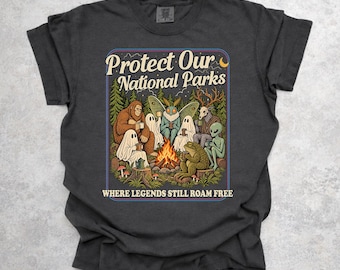 May include: A charcoal gray t-shirt featuring a graphic of Bigfoot, ghosts, a moth, a deer-headed figure, an alien, and a frog sitting around a campfire in a forest. The text reads Protect Our National Parks and WHERE LEGENDS STILL ROAM FREE.