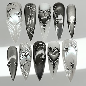 May include: A set of ten long, pointed artificial nails. Each nail features a unique design in shades of silver and black, with abstract patterns, heart shapes, and starburst accents. The nails have a glossy finish.