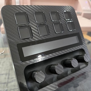 May include: A black carbon fiber textured control deck featuring eight square buttons arranged in two rows, a horizontal rectangular display screen, and four circular knurled knobs positioned along the bottom edge of the device.