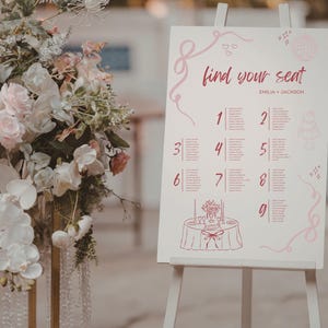May include: A white wedding seating plan on an easel, featuring the words "find your seat" in pink script. The chart displays table numbers and guest names, decorated with pink swirls and illustrations of a cake and table setting.