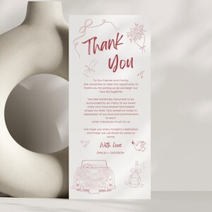 May include: A wedding thank you card with a light pink design. The card features the words "Thank You" in a handwritten font, along with a message and illustrations of a car, flowers, and a dove. The card is vertical and set against a neutral background.
