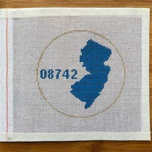 May include: A white needlepoint canvas with a blue silhouette of New Jersey, a gold circle, and the number 08742. The canvas has a red and orange border on the left side and is on a wooden surface.