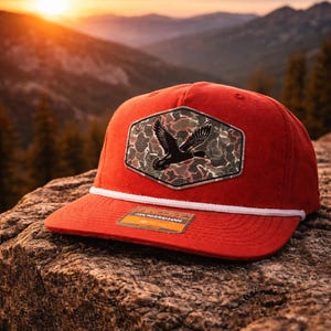 Puede incluir: Gorra de béisbol roja con detalle de cuerda blanca y un parche de camuflaje con un pájaro negro en vuelo. La gorra está sobre un fondo montañoso.