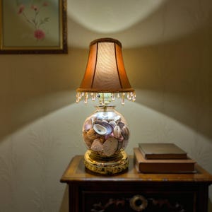 May include: A decorative table lamp with a brown lampshade and a clear glass base filled with seashells. The lamp has a gold-colored base and is illuminated, casting a warm glow. The lampshade is trimmed with a beaded fringe.