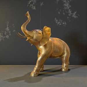 May include: A gold-coloured elephant sculpture with its trunk raised and mouth open. The sculpture has large ears and tusks, and is positioned on a grey surface against a dark grey background. The elephant's shadow is visible.