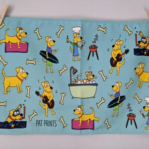 May include: A light blue tea towel with cartoon dogs in various scenes, such as surfing, playing guitar, and barbecuing. The design includes bone and musical note illustrations. The text "PAT PRINTS" is printed on the towel.