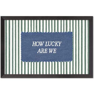 May include: Framed wall art with a striped green and white background. A blue rectangular patch with the text "HOW LUCKY ARE WE" is centered on the artwork. The frame is dark brown.