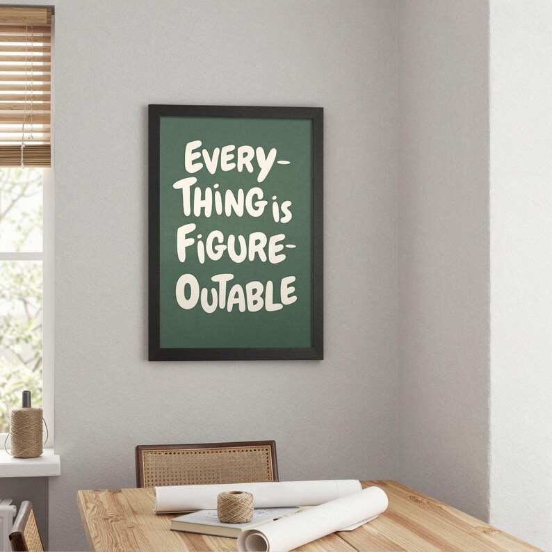 May include: A framed print with the text "EVERYTHING is FIGURE-OUTABLE" in white on a green background. The black frame hangs on a light gray wall. A wooden table and chair are in the foreground.