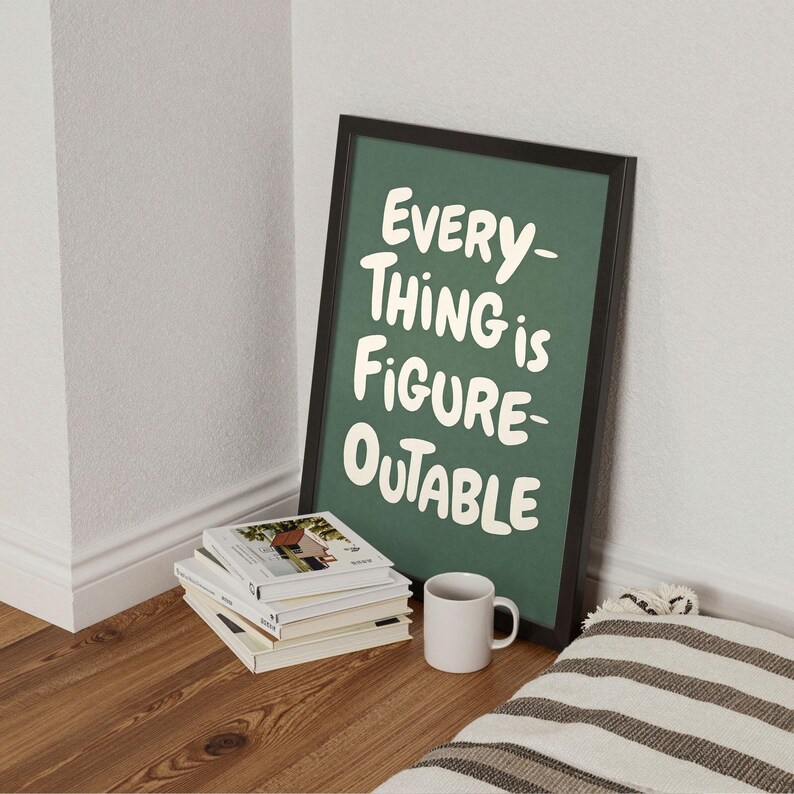 May include: A framed poster with the text "EVERYTHING is FIGURE-OUTABLE" in white on a green background. The poster is propped against a wall next to a stack of books and a white mug on a wooden floor.