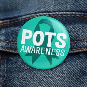 Pode incluir: Um botão redondo turquesa com o texto branco « POTS AWARENESS ». O botão apresenta um design de fita turquesa e está colocado sobre um fundo de ganga. O botão tem um acabamento brilhante.