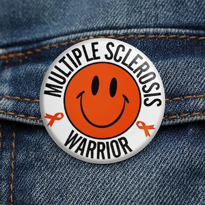 May include: A white button with the words "MULTIPLE SCLEROSIS WARRIOR" in black around the edge. An orange smiley face is in the center. Two orange ribbons are on either side of the smiley face.