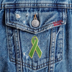 May include: A green awareness ribbon pin with a gold-toned metal border attached to the front pocket of a blue denim jacket. The jacket features a silver metal button and distressed fabric details near the pocket flap and stitching.