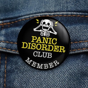 May include: A black circular badge with the words "PANIC DISORDER CLUB MEMBER" in yellow and white. The badge features a white skeleton with its hands on its head and yellow lightning bolts. The badge is set against a blue denim background.