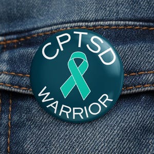 Puede incluir: Un botón redondo de color verde azulado con el texto "CPTSD WARRIOR" en blanco. Una cinta verde azulado está en el centro. El botón está sobre un fondo de mezclilla.