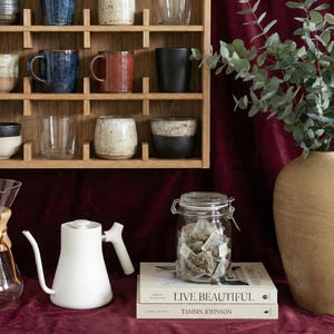 K&ouml;nnte beinhalten: Ein wei&szlig;er Wasserkocher mit langem Ausguss und ein Holzregal mit verschiedenen Tassen und Gl&auml;sern. Ein Glas mit Teebeuteln steht auf zwei B&uuml;chern mit dem Titel "LIVE BEAUTIFUL". Rechts steht eine gro&szlig;e Vase mit gr&uuml;nen Bl&auml;ttern.