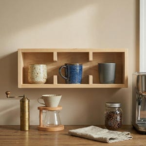 K&ouml;nnte beinhalten: Ein Holzregal zeigt drei Keramikbecher in Beige, Blau und Grau. Eine manuelle Kaffeem&uuml;hle, ein Pour-Over-Kaffeebereiter und ein Glas Kaffeebohnen stehen auf der Holzoberfl&auml;che darunter. Ein gefaltetes Leinentuch liegt ebenfalls auf der Oberfl&auml;che.