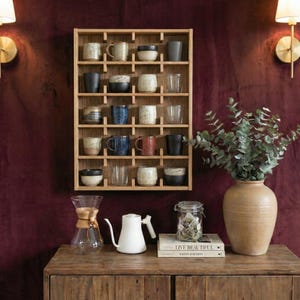 May include: A wooden display shelf with an assortment of ceramic mugs and bowls. A glass carafe, white kettle, and a jar with a book are on a wooden table. A large vase with greenery sits on the table, with a burgundy wall in the background.