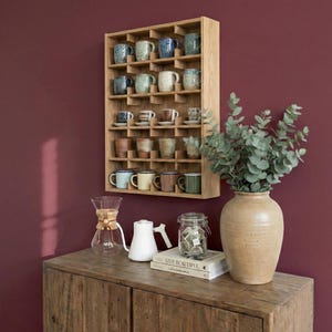 K&ouml;nnte beinhalten: Ein h&ouml;lzernes Tassenregal mit mehreren F&auml;chern, gef&uuml;llt mit verschiedenen Keramiktassen in Blau-, Gr&uuml;n- und Braunt&ouml;nen. Das Regal ist an einer tiefroten Wand befestigt. Ein Holzschrank mit einer Vase, einer Glaskaraffe und einem wei&szlig;en Wasserkocher befindet sich im Vordergrund.
