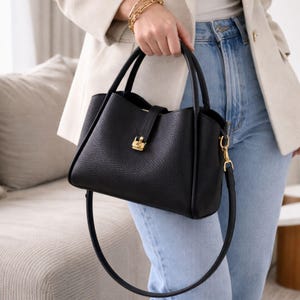 May include: A black leather handbag with a textured finish. The bag has a structured shape with a top handle and a detachable shoulder strap. It features gold-coloured hardware, including a clasp closure and strap attachments. The bag is held by a person wearing a cream-coloured blazer and blue jeans.