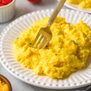 May include: A plate of scrambled eggs, bright yellow in colour, with a silver fork inserted. The white plate has a decorative rim. Small green herbs are sprinkled on top of the eggs. The eggs appear fluffy.