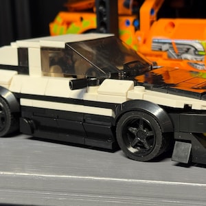 May include: A white and black toy car with black wheels. The car has a spoiler and a black front bumper. The car is a model of a sports car. The car is made of plastic.