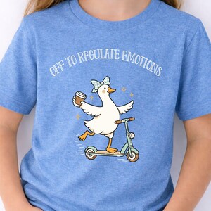 May include: A light blue t-shirt with a cartoon duck design. The duck, wearing a bow, rides a scooter and holds a coffee cup. The text "OFF TO REGULATE EMOTIONS" is printed in white on the shirt.