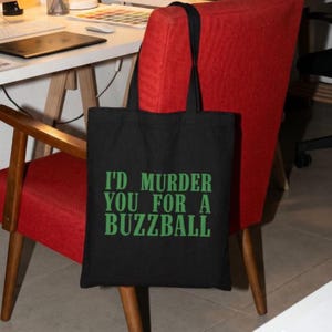 May include: Black tote bag with the text "I'D MURDER YOU FOR A BUZZBALL" in green. The bag has black handles and is hanging on a red chair. The bag is made of a sturdy material and is suitable for carrying groceries or other items.