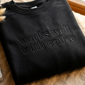 May include: Black crew neck sweatshirt with the phrase "works well with coffee" embroidered on the front. The sweatshirt is displayed on a beige surface near a window, next to a notebook and dried flowers.