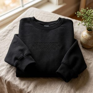 May include: Black crew neck sweatshirt with the embroidered text "MY BODY HURTS AND I'M MAD AT THE GOVERNMENT". The sweatshirt is folded on a neutral-coloured surface.
