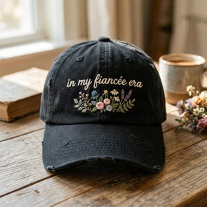 Puede incluir: Gorra de béisbol negra con detalles desgastados. La gorra presenta la frase bordada "in my fiancée era" sobre un diseño floral con flores rosas, azules y amarillas. La gorra está sobre una superficie de madera.