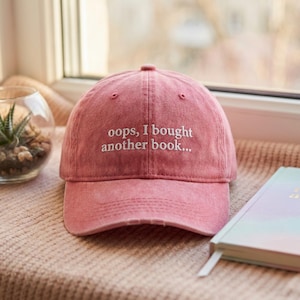 May include: A faded pink baseball cap with the embroidered text "oops, I bought another book...". The cap is on a pink surface next to a book and a small glass terrarium with a succulent plant.