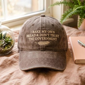 Może przedstawiać: Brązowa czapka bejsbolowa z haftowanym napisem "I BAKE MY OWN BREAD & DON'T TRUST THE GOVERNMENT" i wzorem kłosa pszenicy. Czapka wykonana jest z zużytej, teksturowanej tkaniny.