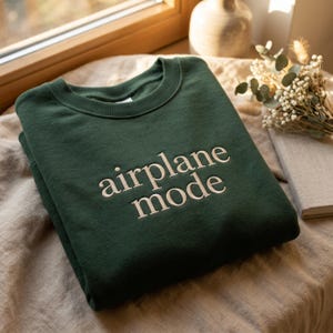 May include: A dark green sweatshirt with the words "airplane mode" embroidered in a light beige color. The sweatshirt is folded and placed on a neutral-toned surface, with a window and decorative items in the background.