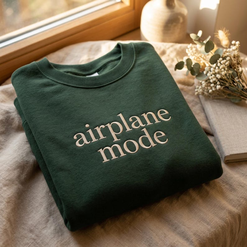 May include: A dark green sweatshirt with the words "airplane mode" embroidered in a light beige color. The sweatshirt is folded and placed on a neutral-toned surface, with a window and decorative items in the background.