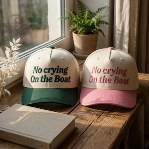 May include: Two trucker hats with off-white crowns and mesh backs. One hat features a dark green brim and dark green embroidered text that reads No crying On the Boat. The other hat has a pink brim and pink embroidered text reading No crying On the Boat.