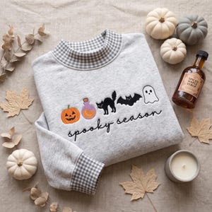 May include: A light gray heathered sweatshirt with a gingham collar and cuffs. The front features embroidered icons of a pumpkin, potion bottle, black cat, bat, and ghost, with the black cursive text "spooky season" embroidered below.