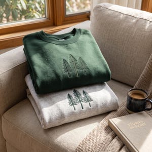 May include: A dark forest green crewneck sweatshirt stacked on top of a light heather gray crewneck sweatshirt. Both garments feature an identical embroidered design of three pine trees in dark green thread centered on the chest area.