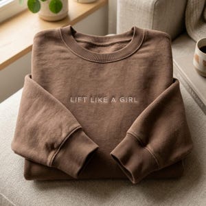 May include: A folded brown sweatshirt with the embroidered text "LIFT LIKE A GIRL" in white. The sweatshirt has a crew neck and ribbed cuffs. The image is taken from a slightly elevated angle.